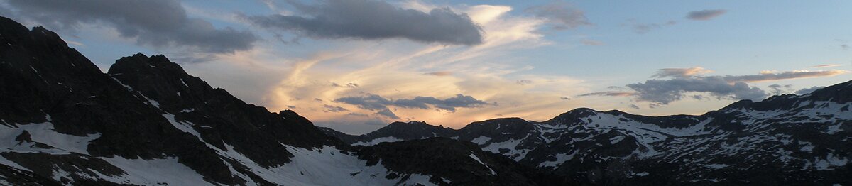 Veduta delle Alpi Marittime in tarda primavera, con le cime ancora innevate illuminate dalla luce del tramonto e il cielo attraversato da nuvole dorate. Archivio APAM. Fine descrizione dell’immagine.