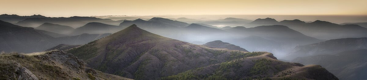 Veduta panoramica della Cima Gouron, con la luce dorata che sfuma sulle creste montuose e le valli immerse in una leggera foschia. Archivio APAM. Fine descrizione dell’immagine.