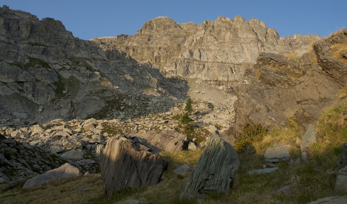 Veduta del versante occidentale del Monte Bego nella Valle Roya, con pareti rocciose illuminate dalla luce del pomeriggio e massi sparsi tra prati e pietraie. Foto di G. Bernardi. Fine descrizione dell’immagine.